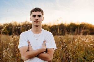 Person in white t-shirt standing with crossed arms in golden field at sunset with trees in background.