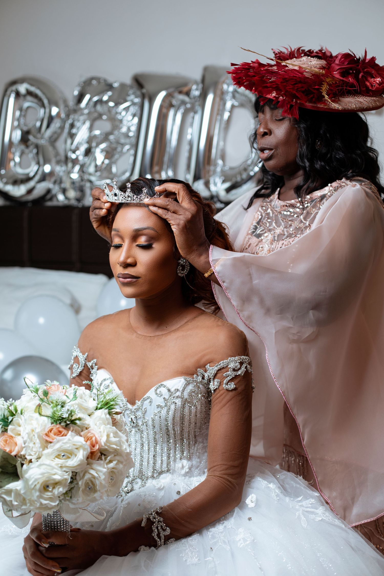 A bride in an ornate white wedding dress with lace details holds a bouquet while having a tiara placed on her head.
