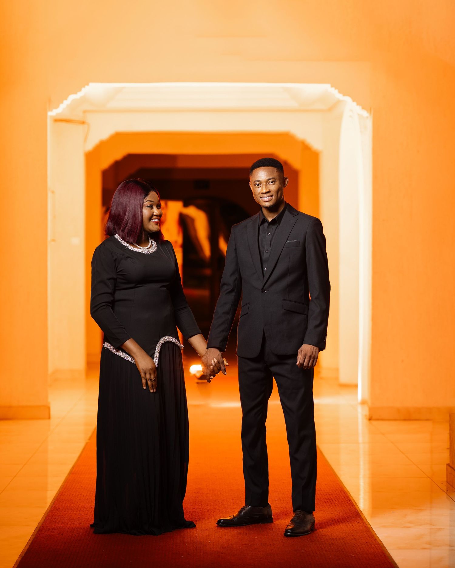 A couple poses in formal black attire against an orange-lit archway backdrop.