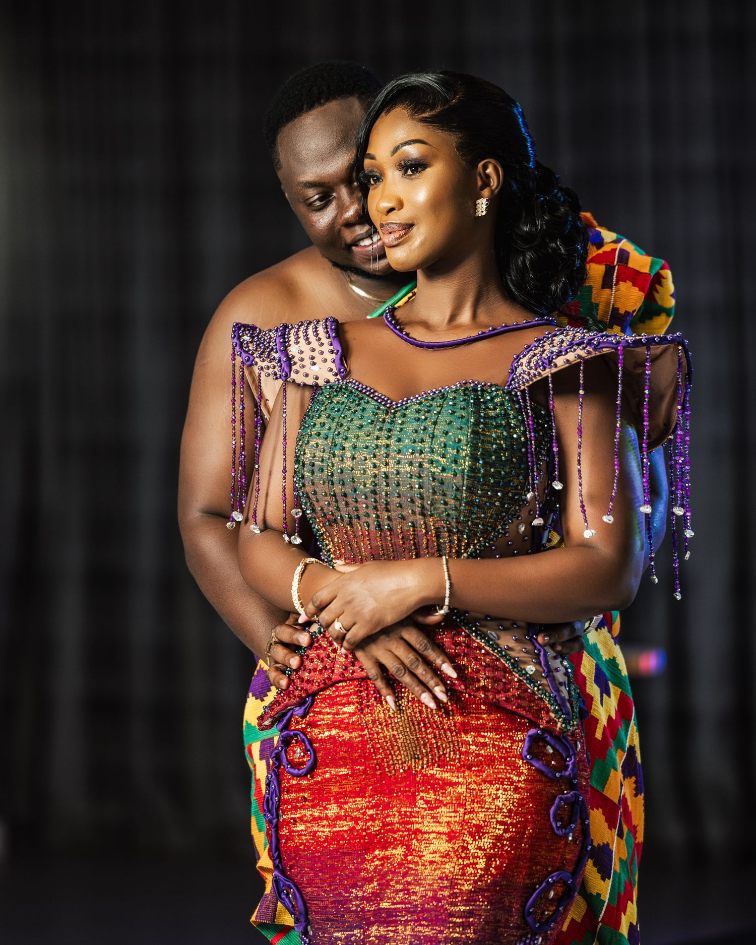 A couple embraces in colorful traditional African attire with beaded details and vibrant patterns.