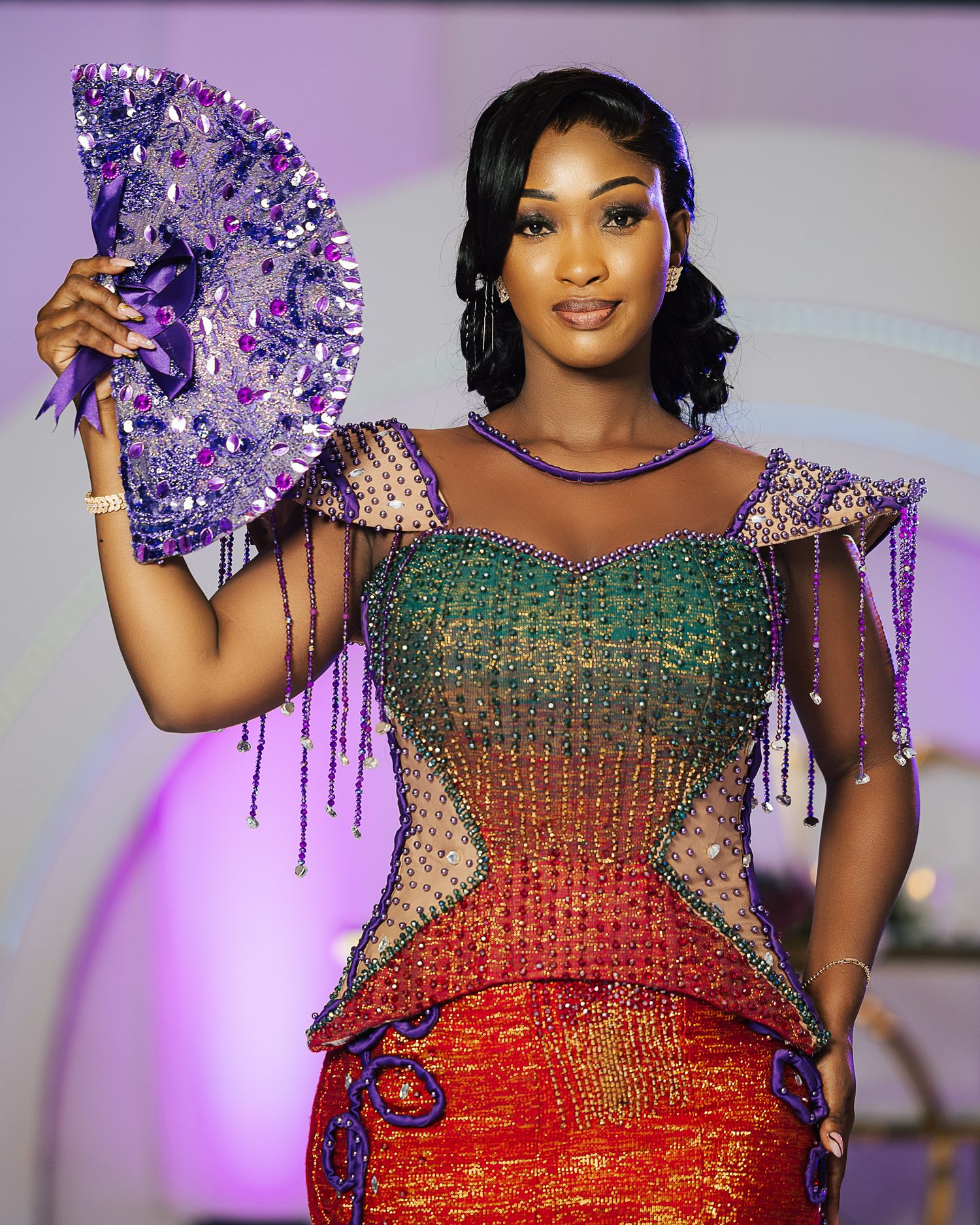 A model poses with a purple fan wearing a stunning red and green beaded dress with sheer details.