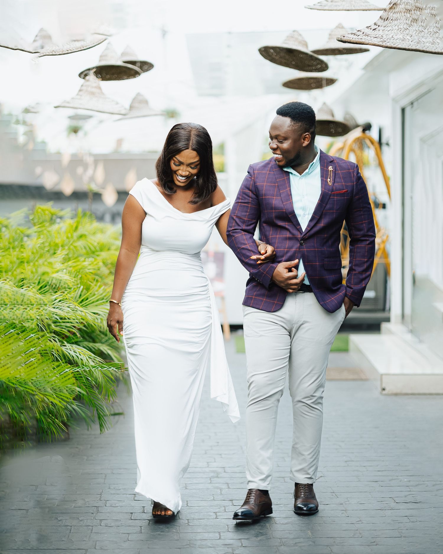 A couple walks together on a modern pathway wearing coordinated white and purple formal outfits.