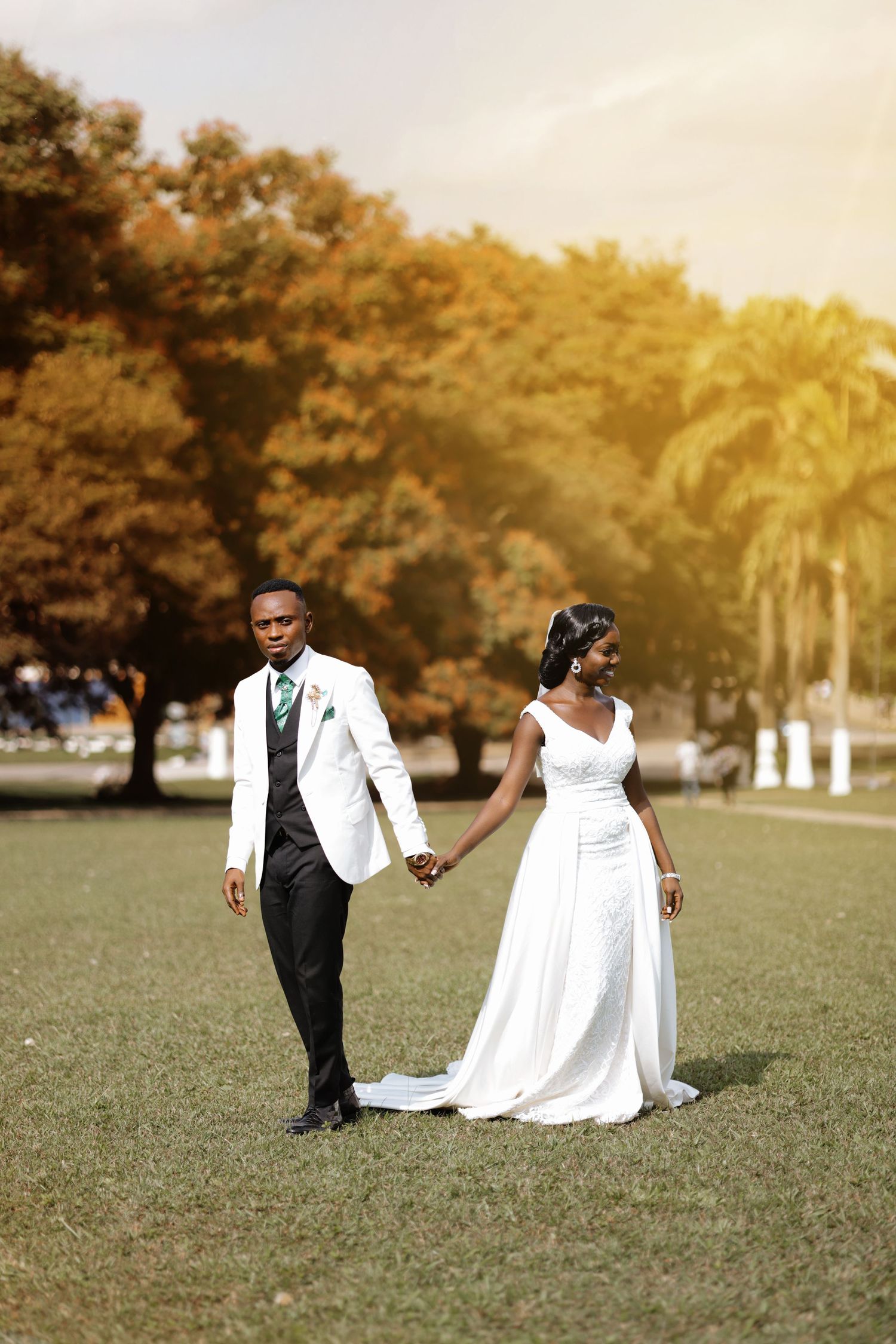 A romantic wedding photoshoot in a park with a flowing white dress and tuxedo against a golden sunset background.