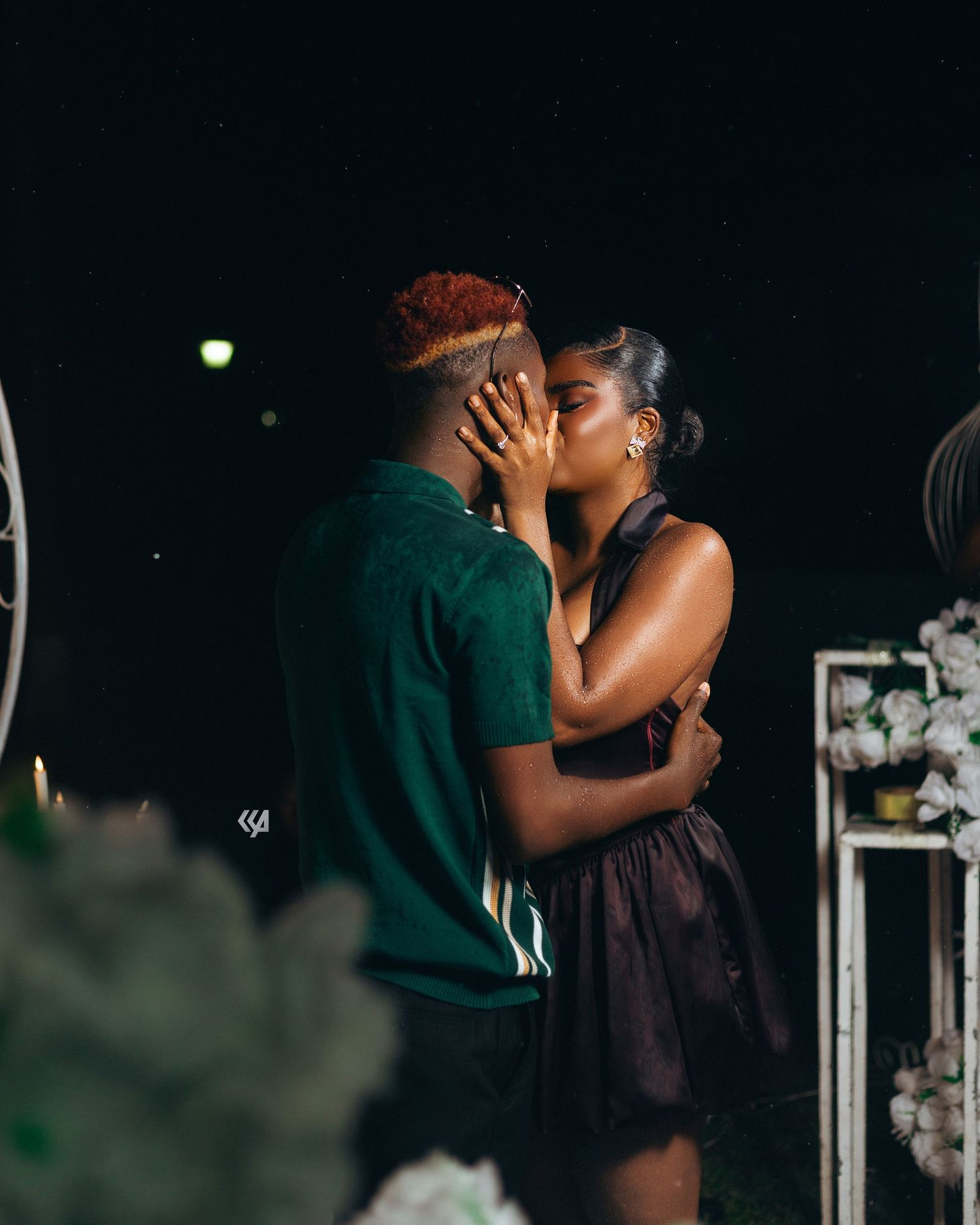Couple sharing a romantic kiss at night near white floral decorations in an outdoor setting.