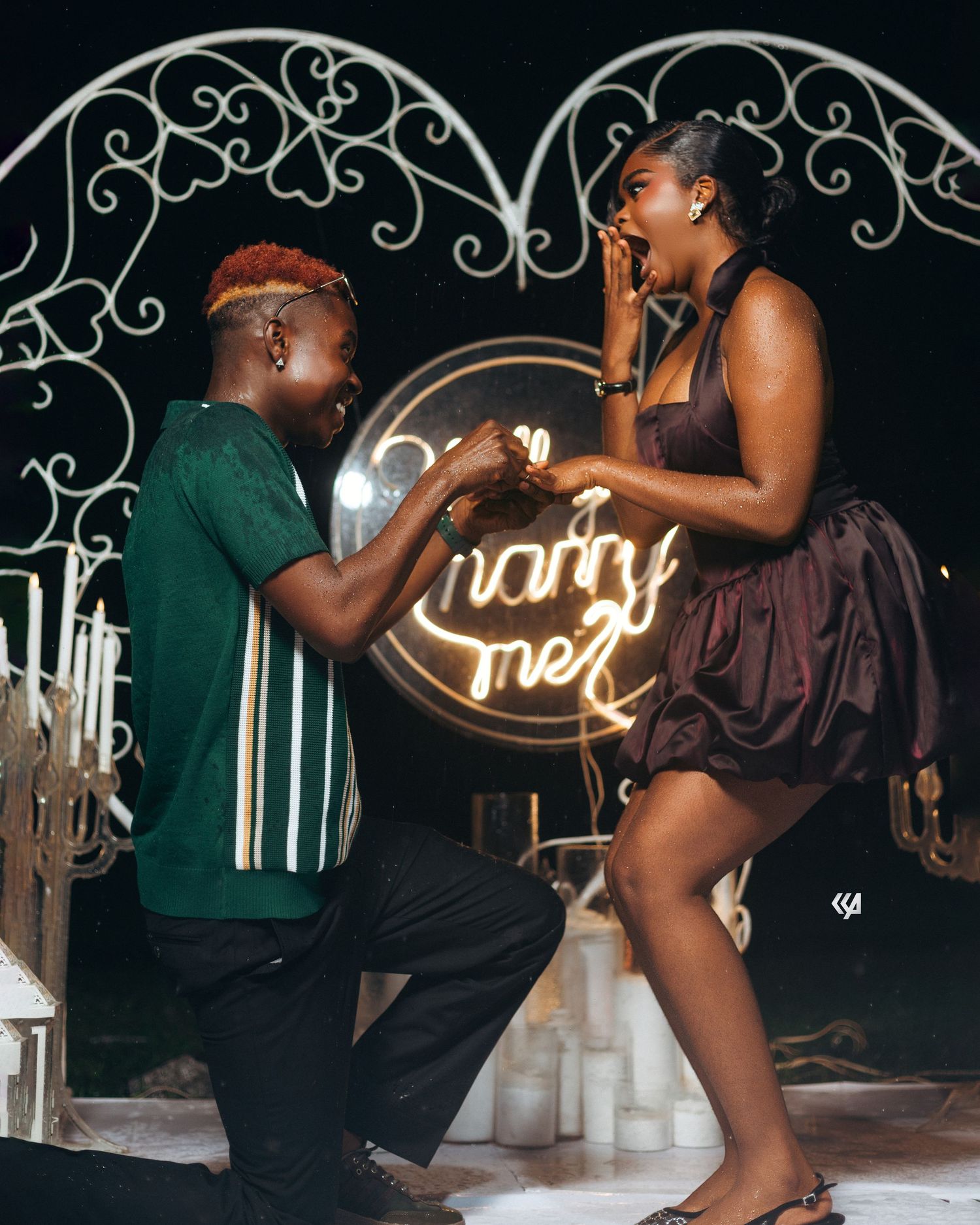 A man kneels on one knee proposing to a surprised woman in front of a glowing marry me sign at night.