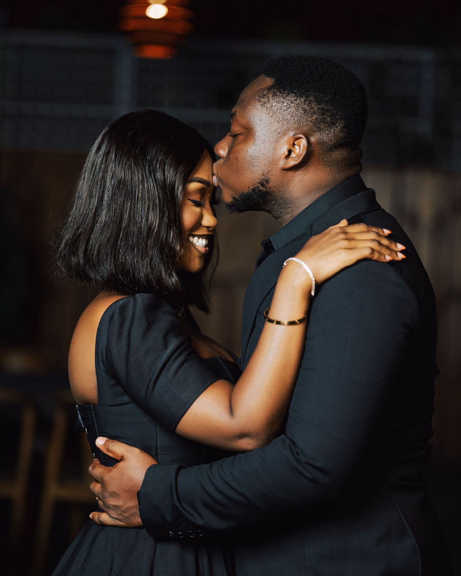 A romantic couple embraces intimately in a dimly lit setting wearing dark formal attire.
