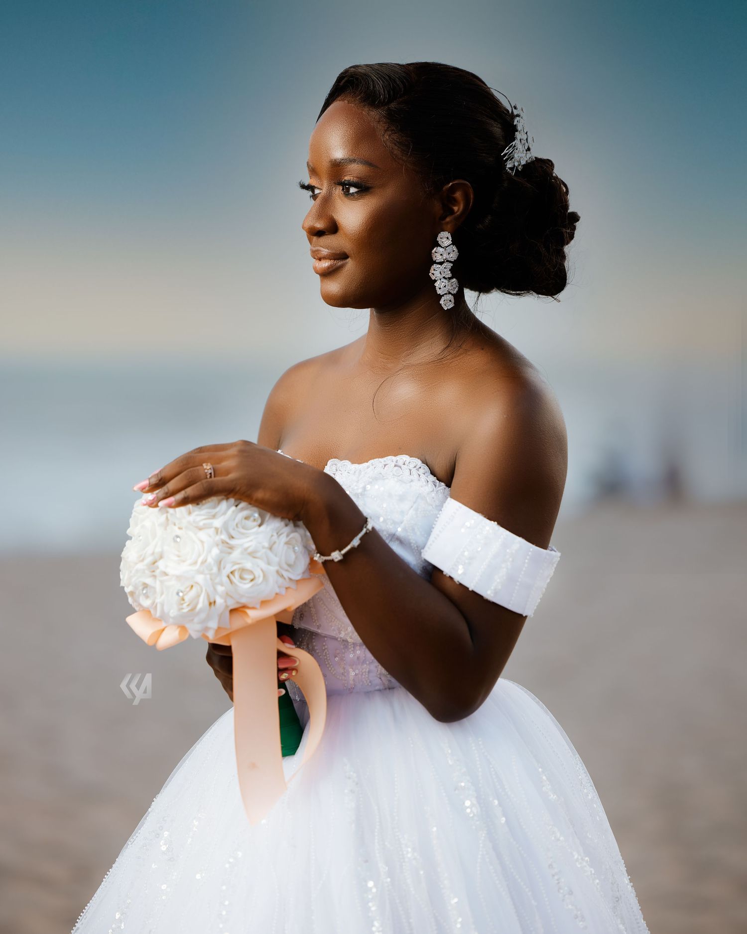 A bride in an off-shoulder white wedding gown poses with her bouquet against a sunset backdrop.
