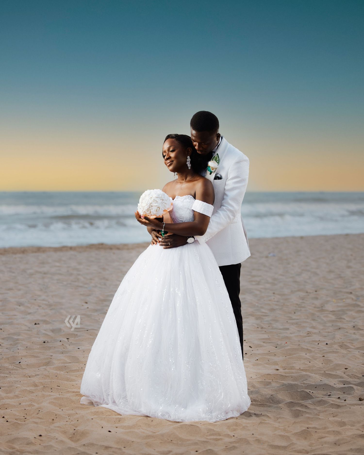 A romantic beach wedding photo of a couple embracing at sunset in formal attire.
