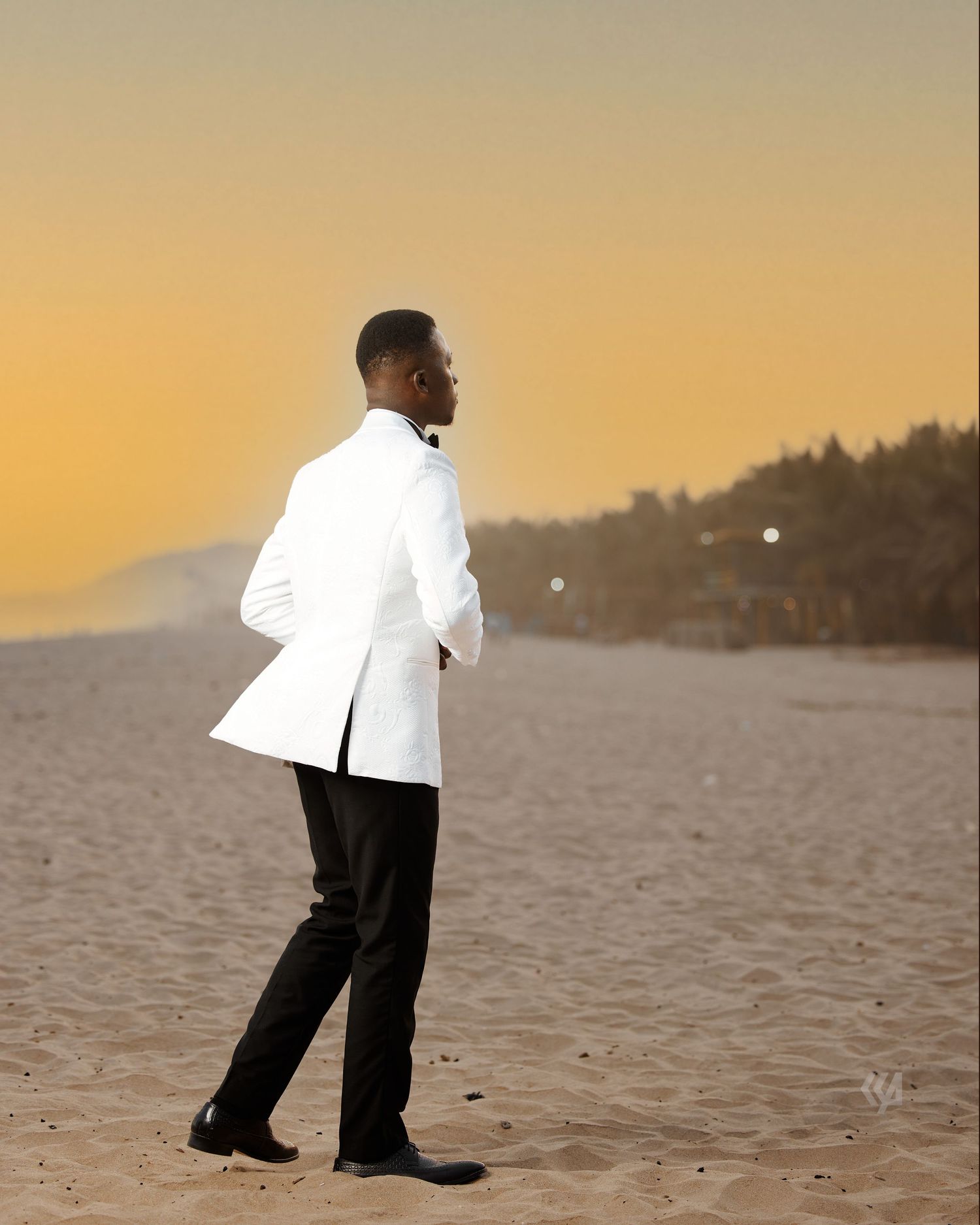 A groom in a white tuxedo stands on a beach at sunset.