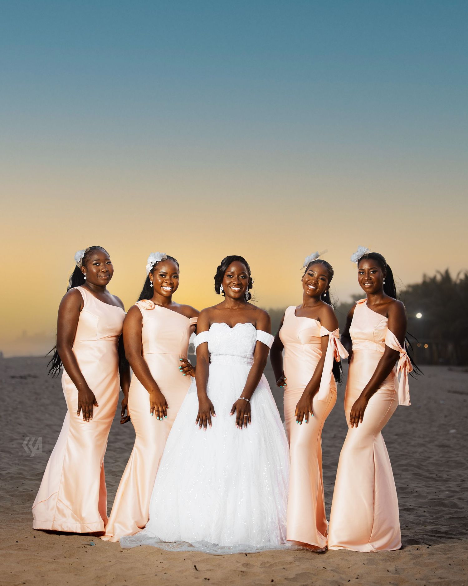 A bridal party in blush and white dresses poses on the beach at sunset.