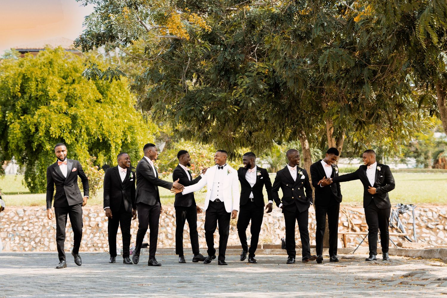 A wedding party in formal attire poses together under trees.