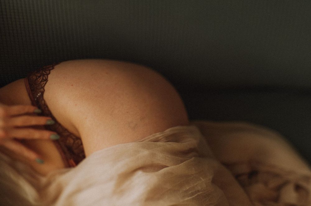 Intimate boudoir photo showing bare shoulder and delicate lace lingerie against soft white bedding in moody lighting.