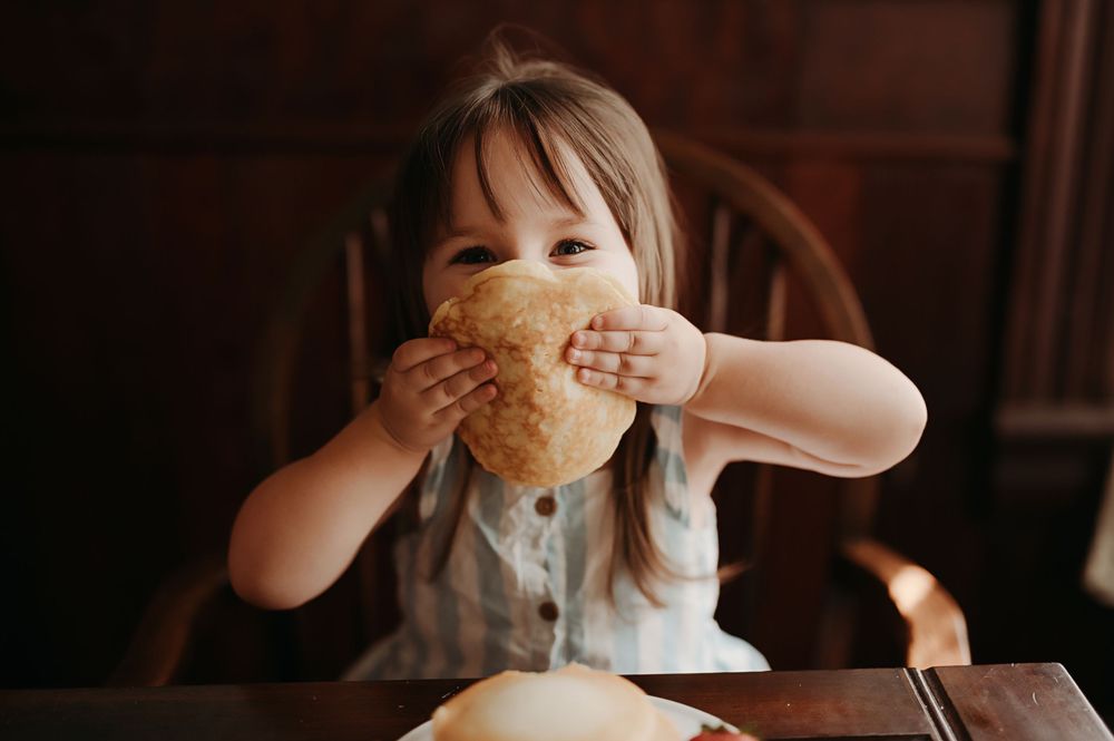 A young child in a white dress sits at a table eating a pastry in a dimly lit room.