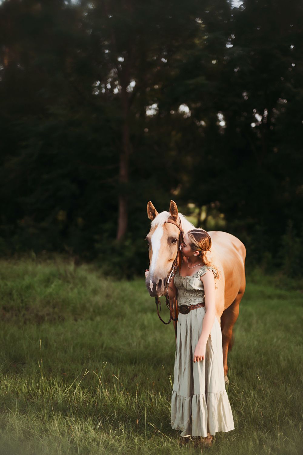 A palomino horse stands beside someone wearing a flowing white dress in a grassy field at dusk.