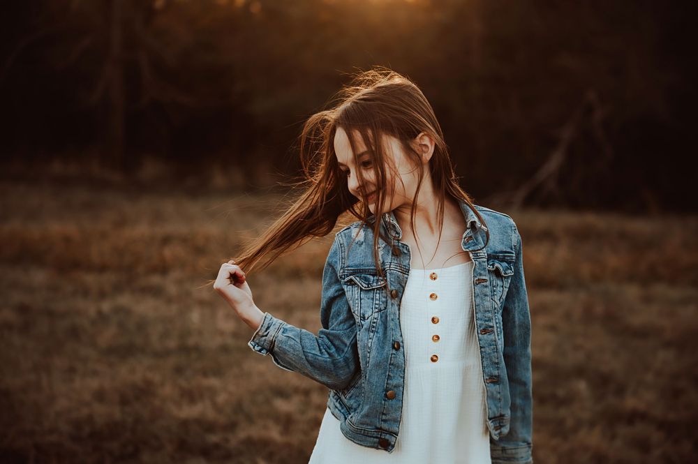 A young girl in a denim jacket and white shirt stands in a rustic outdoor setting during golden hour.