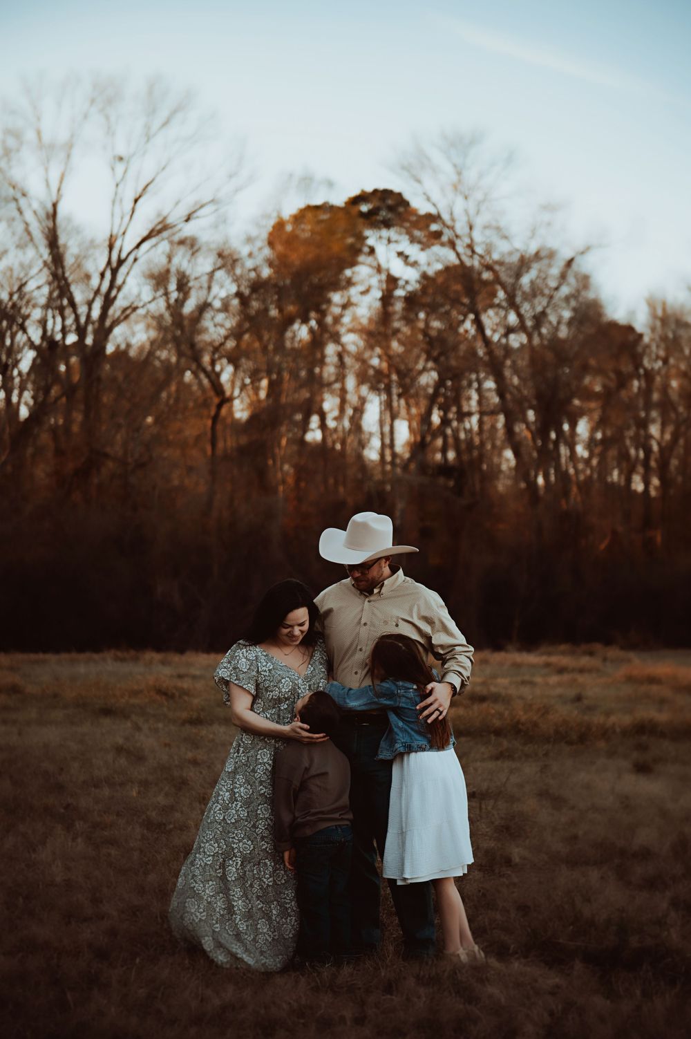 A family shares tender moments together in a rustic field during golden hour as autumn trees line the background.