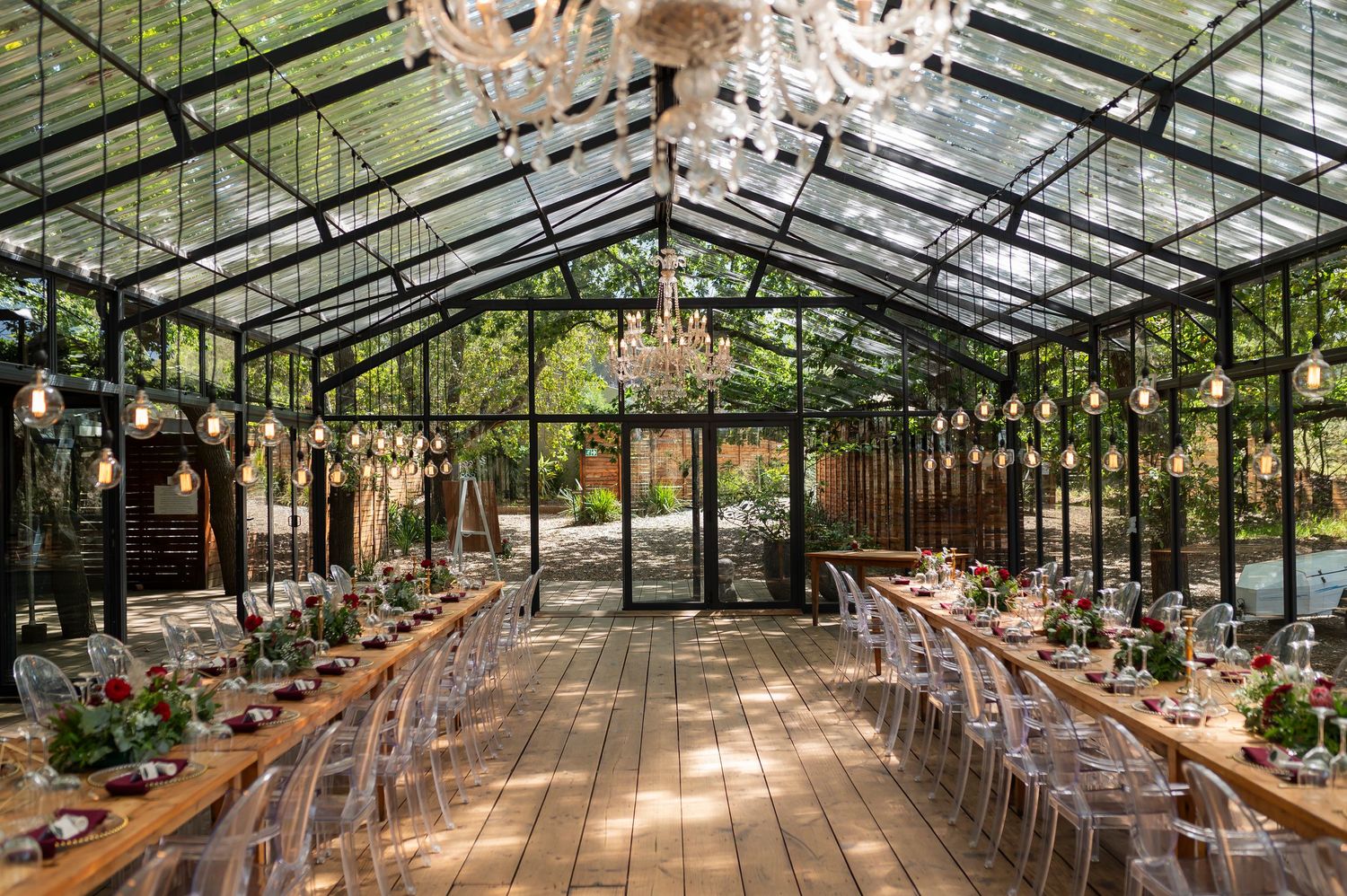 Long wooden banquet tables arranged in rows under a glass greenhouse ceiling with hanging string lights and greenery.
