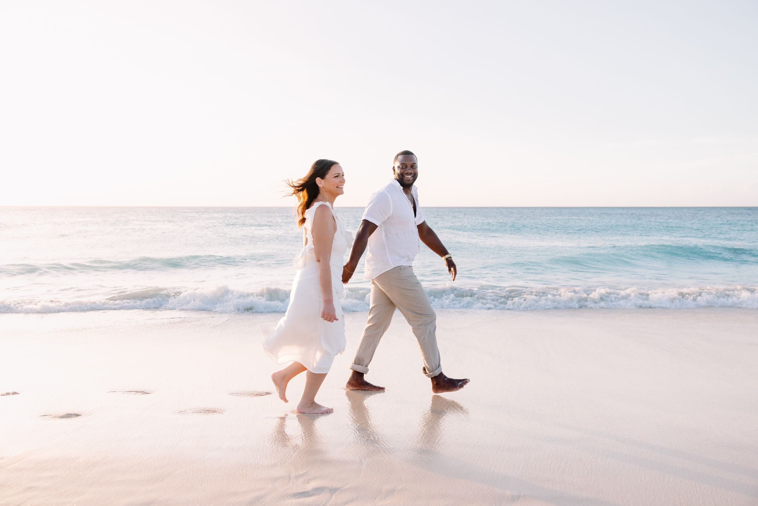 Couple walking hand in hand on a sunny beach with white sand and blue ocean waves.