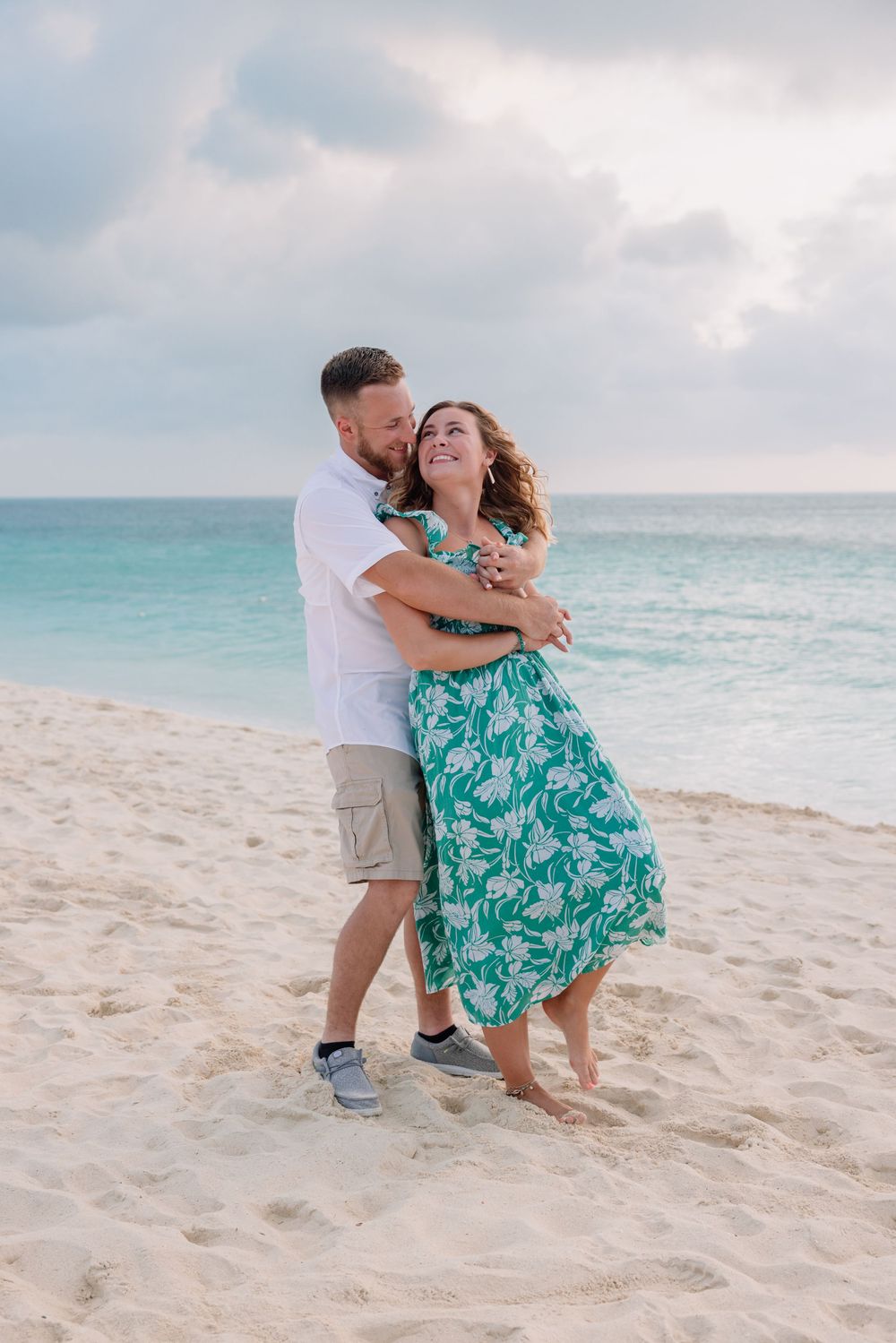 Couple embracing on a white sandy beach with turquoise ocean, woman in green floral dress, man in white shirt and khaki shorts.