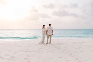 Couple walking along empty beach with gentle waves and pastel sunset sky.