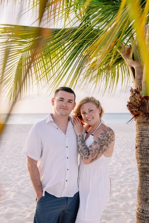 Couple in matching white outfits posing under swaying palm trees on a sandy beach.
