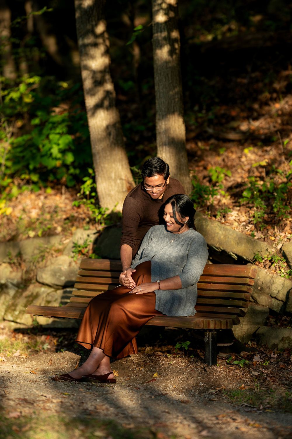 Expectant couple sitting on park bench in autumn forest, man embracing pregnant woman as she looks down at her belly.
