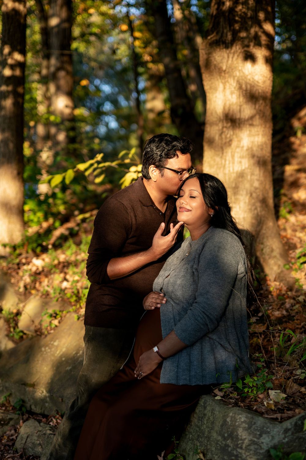 Couple in autumn forest, man kissing pregnant woman's cheek while gently holding her baby bump against rocky backdrop.