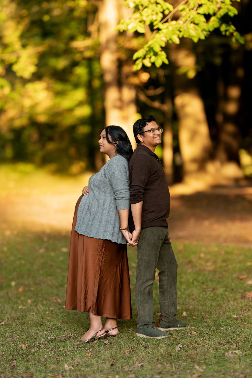 Pregnant couple standing back-to-back holding hands in autumn park, woman cradling baby bump in gray sweater and brown skirt.