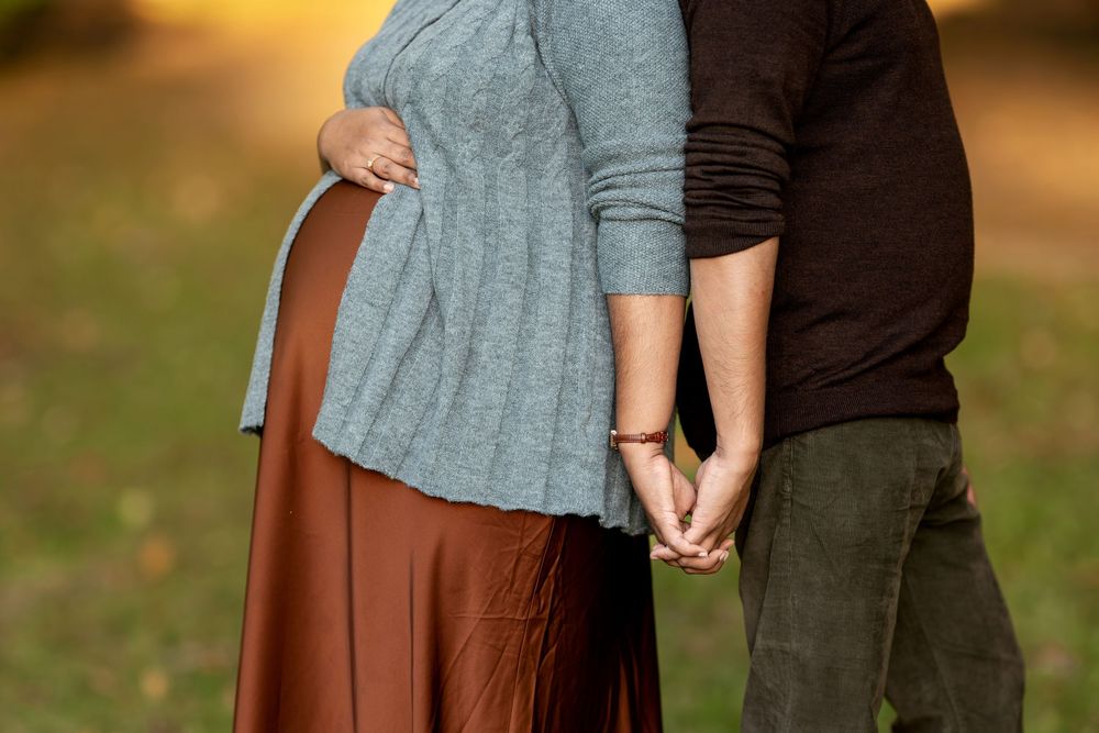 Close-up of pregnant woman's baby bump in gray sweater and rust skirt, partner holding hands beside her outdoors.