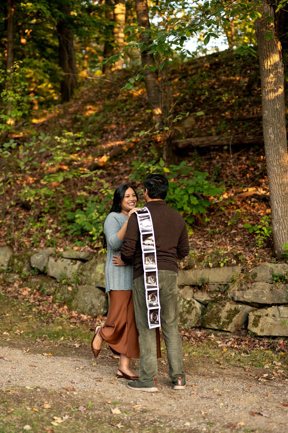 Expecting couple embracing in autumn forest while man holds a long strip of ultrasound photos behind his back.