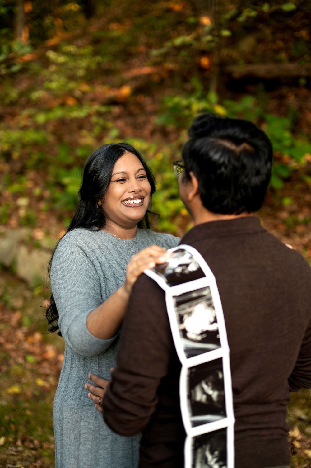 Joyful pregnant woman laughing with partner holding ultrasound strip in vibrant autumn forest setting.