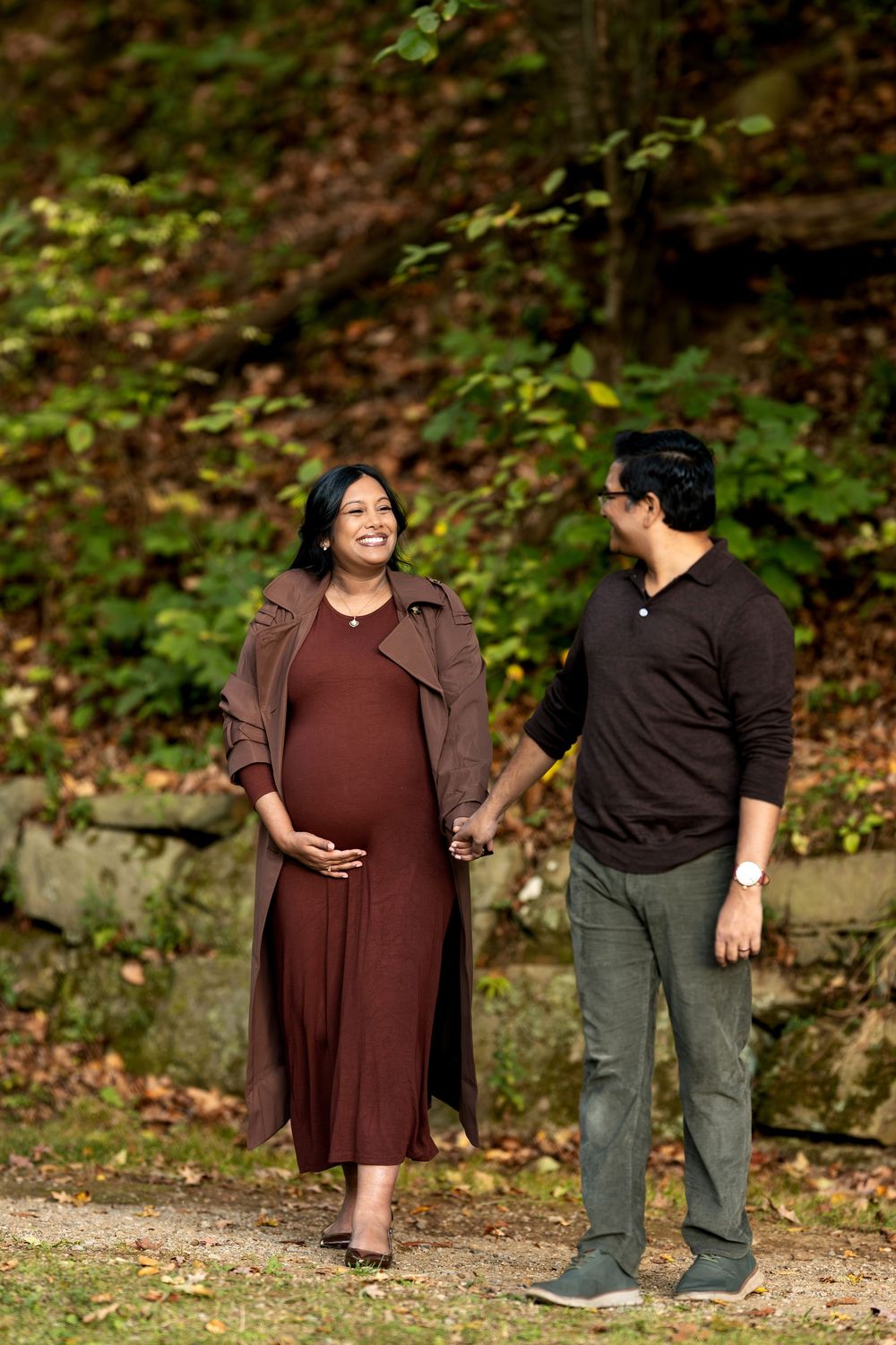 Happy pregnant couple holding hands and laughing together on a scenic fall nature path with lush greenery.