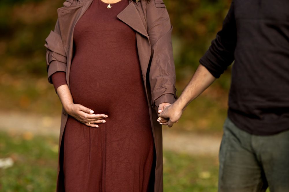 Close-up of pregnant woman in burgundy dress holding baby bump while holding partners hand outdoors.