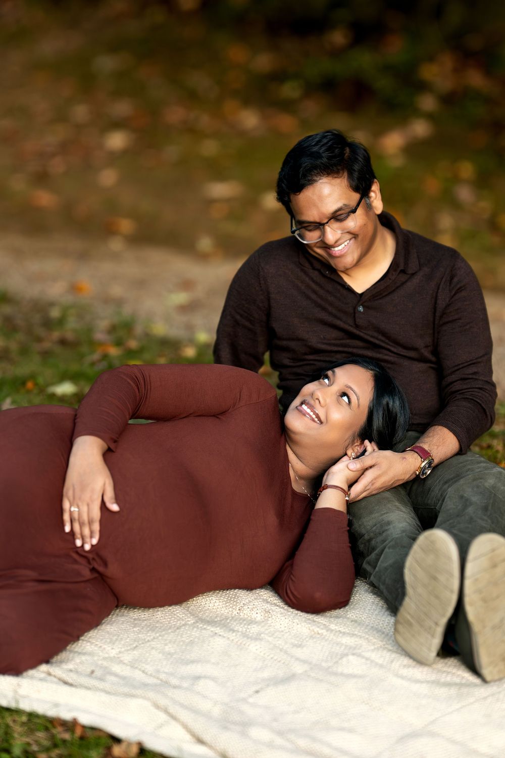 Pregnant woman lying on blanket smiling up at partner in warm autumn park maternity photo session.