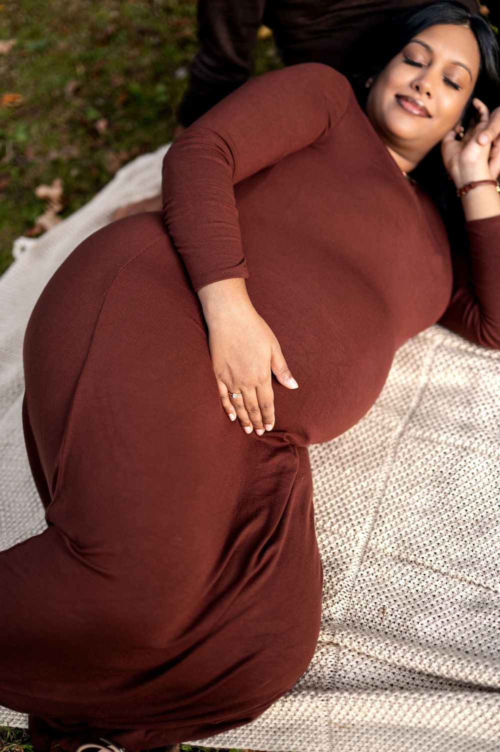 Close-up of pregnant woman in rust-colored dress lying peacefully on blanket outdoors in natural light.