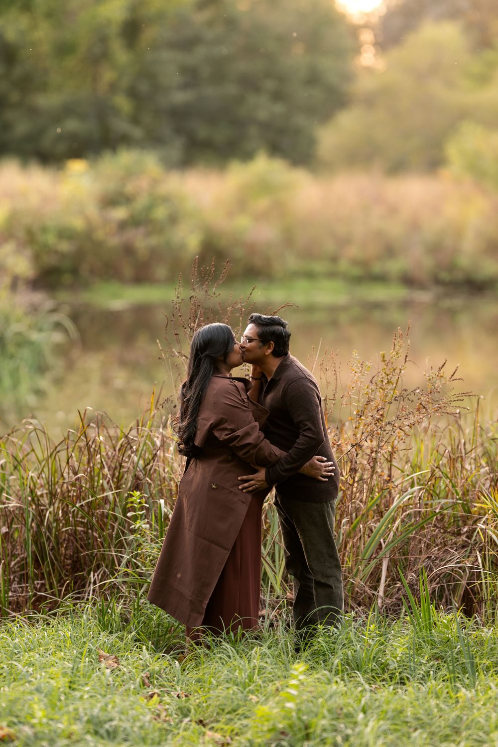 Couple sharing a tender kiss near a tranquil pond with warm autumn grasses and trees in the background.