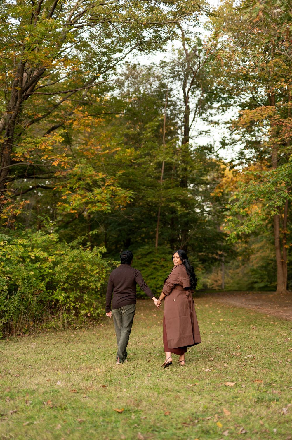 Pregnant couple walking hand in hand through a lush park with vibrant autumn foliage overhead.