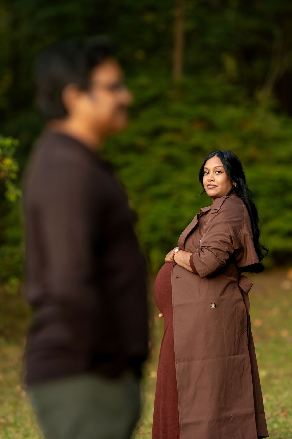 Smiling pregnant woman in brown coat cradling baby bump with partner blurred in foreground in a park.