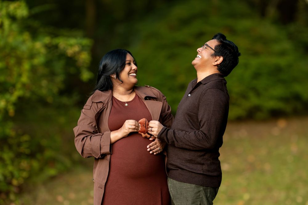 Joyful pregnant couple laughing together outdoors in a green park during a maternity photo session.