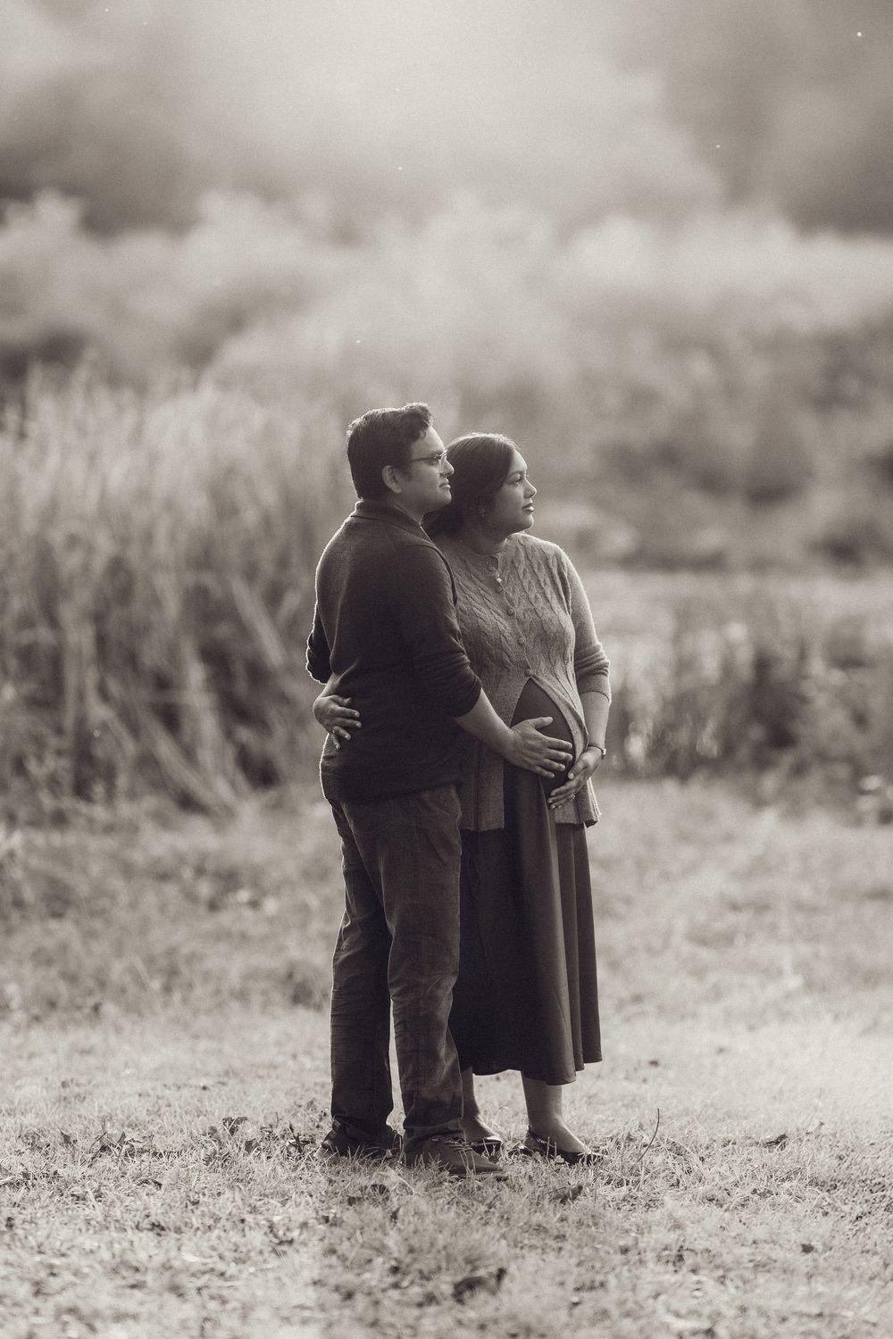 Black and white maternity portrait of couple embracing on a path surrounded by tall grass.
