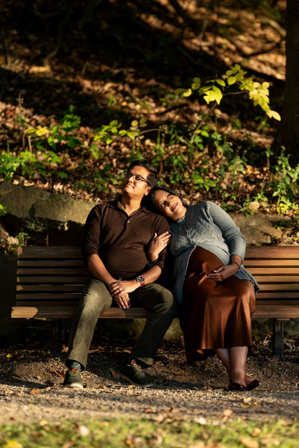 Expectant couple resting on a park bench, woman leaning on man's shoulder, bathed in warm light.