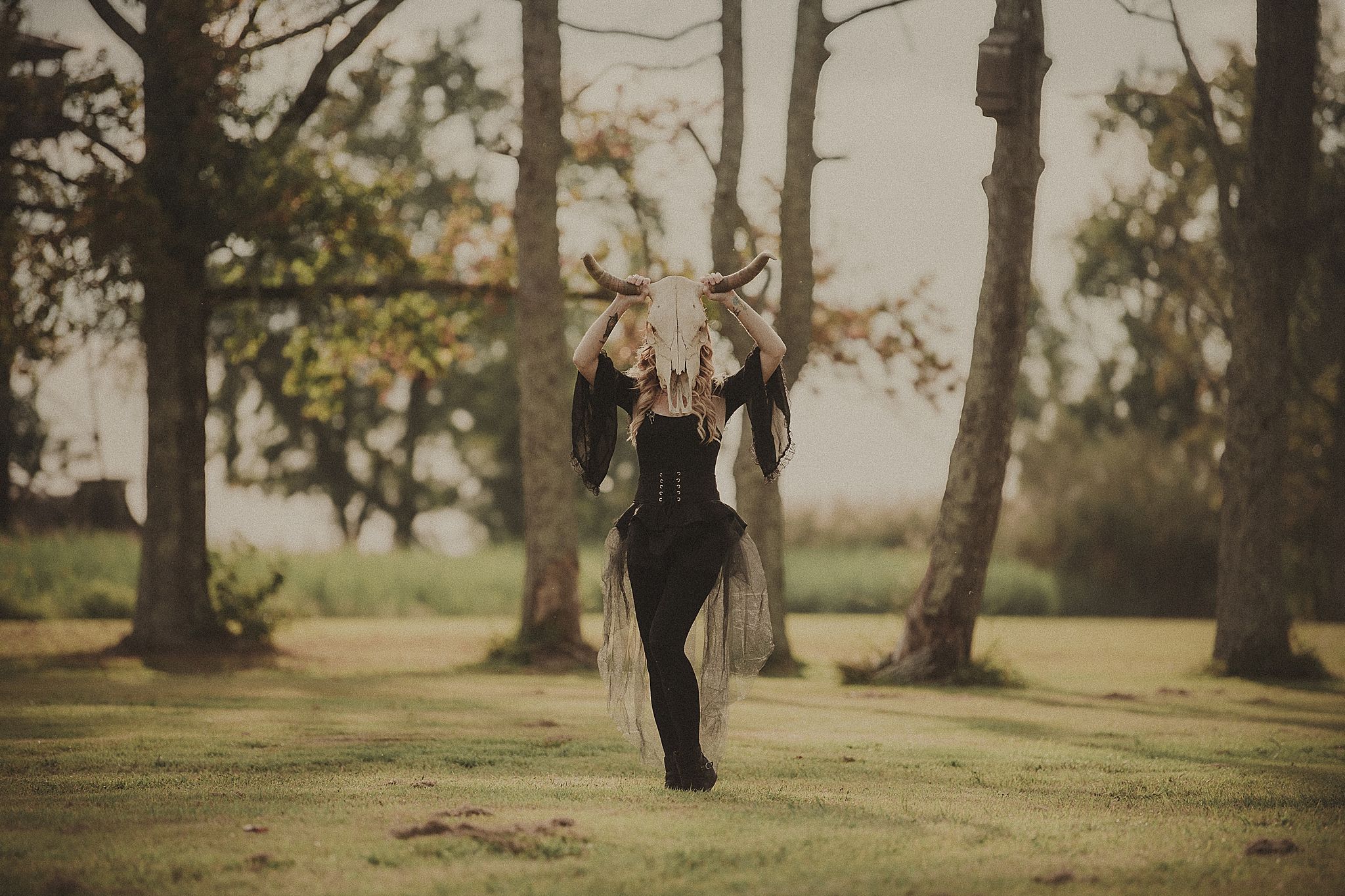 A person in black boots walks across a sunlit grassy park with tall trees casting long shadows in autumn.
