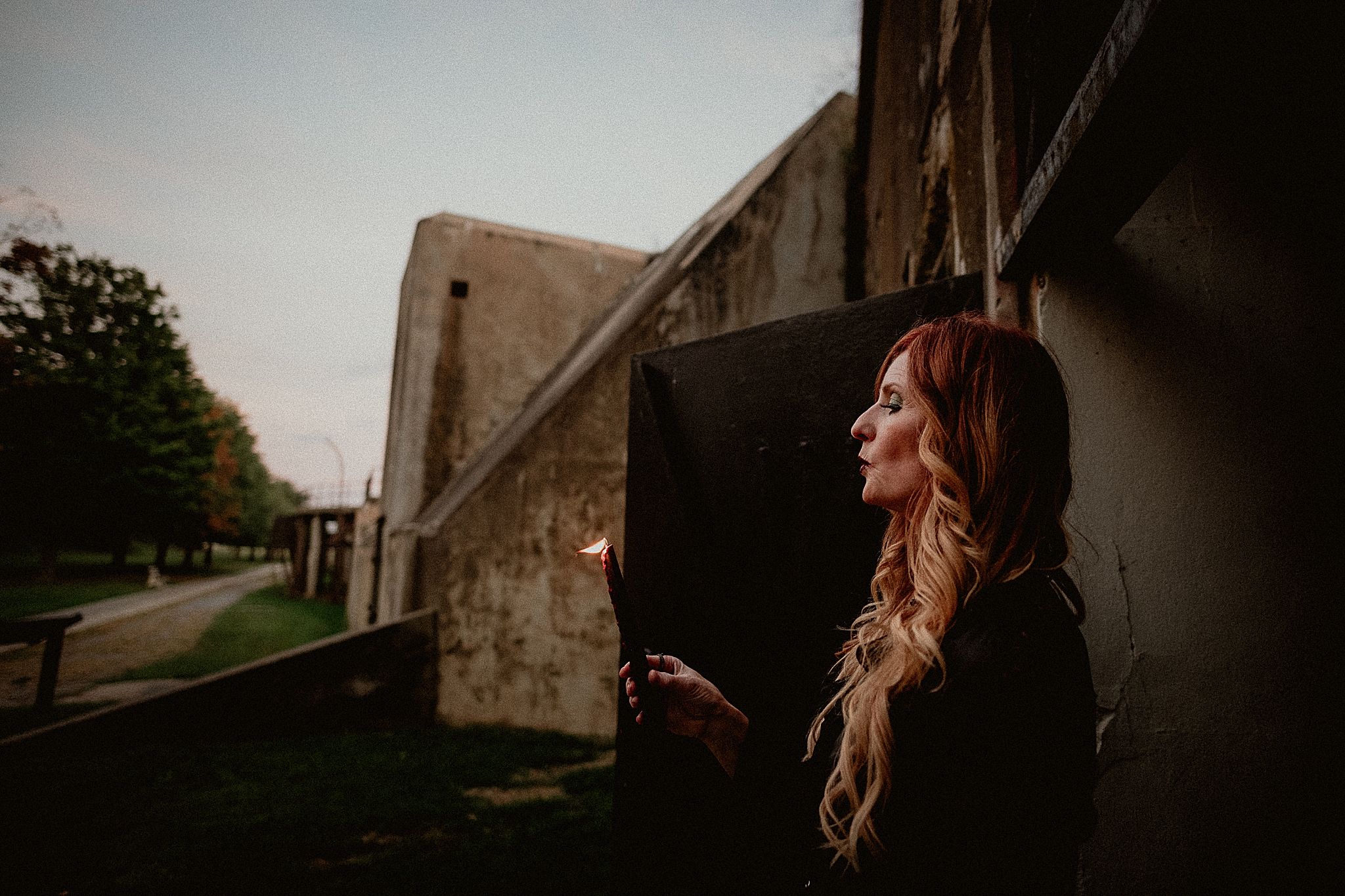 A person with long wavy hair stands in a dramatic pose against a rustic stone wall in warm sunlight.