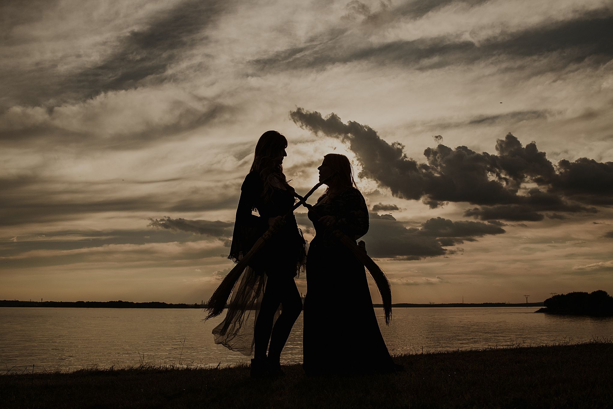Silhouetted figures stand together against a dramatic sunset sky over a tranquil waterfront landscape.