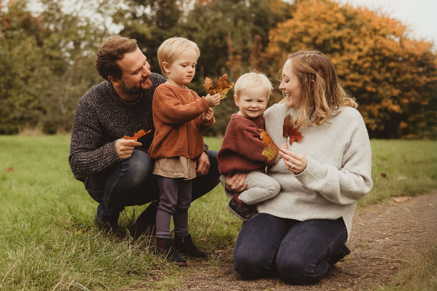 A family sits together on a dirt path surrounded by autumn trees with golden foliage in the background.