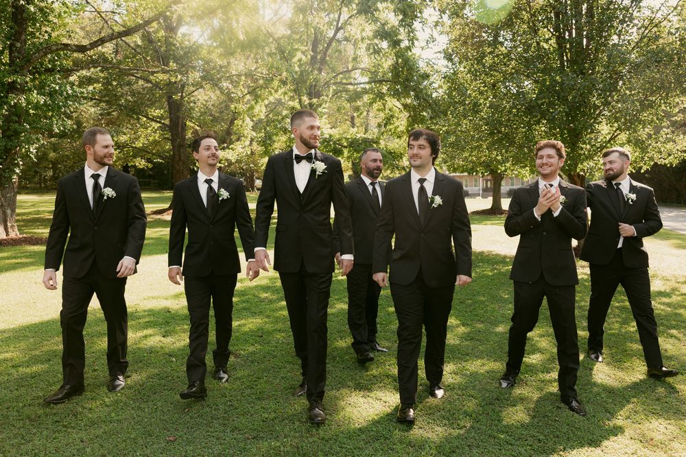 Groomsmen in black tuxedos walk together through a sunlit backyard during a wedding photo session.
