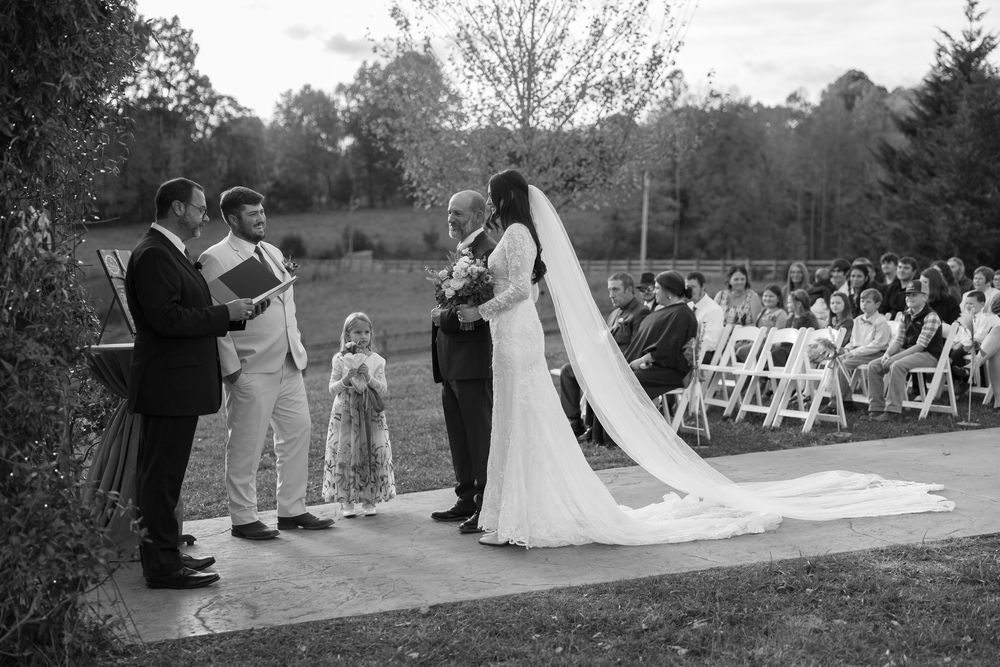 A wedding ceremony takes place outdoors with white chairs arranged on a lawn and a long flowing veil in view.
