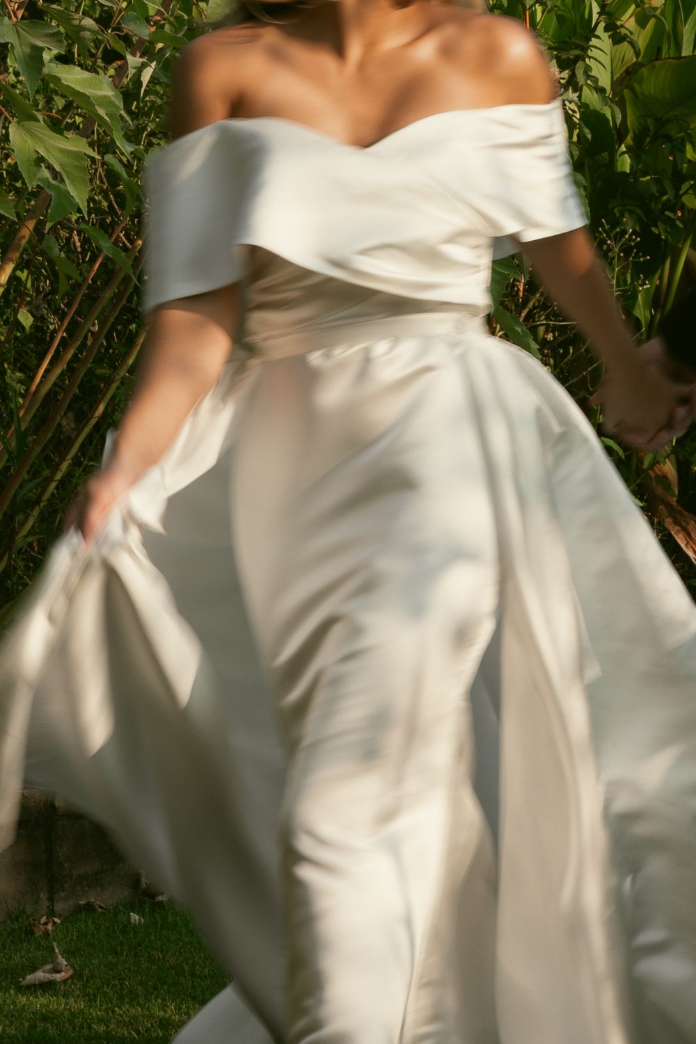 Elegant white wedding dress with off-shoulder neckline and flowing fabric details captured in a garden setting.