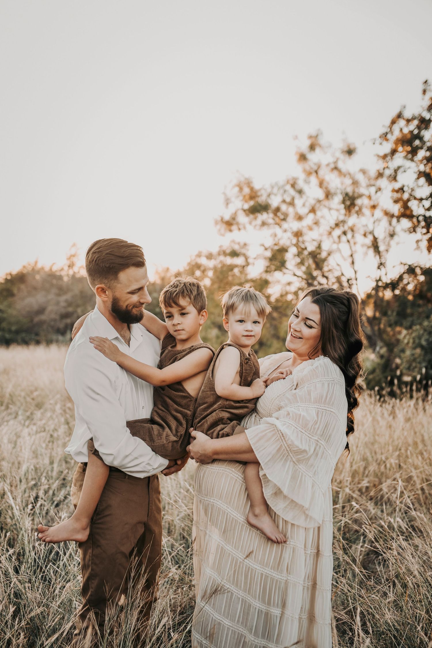 two sweet boys in brown rustic overalls lean back to back as their mom and dad snuggle them and laugh during their family maternity photos