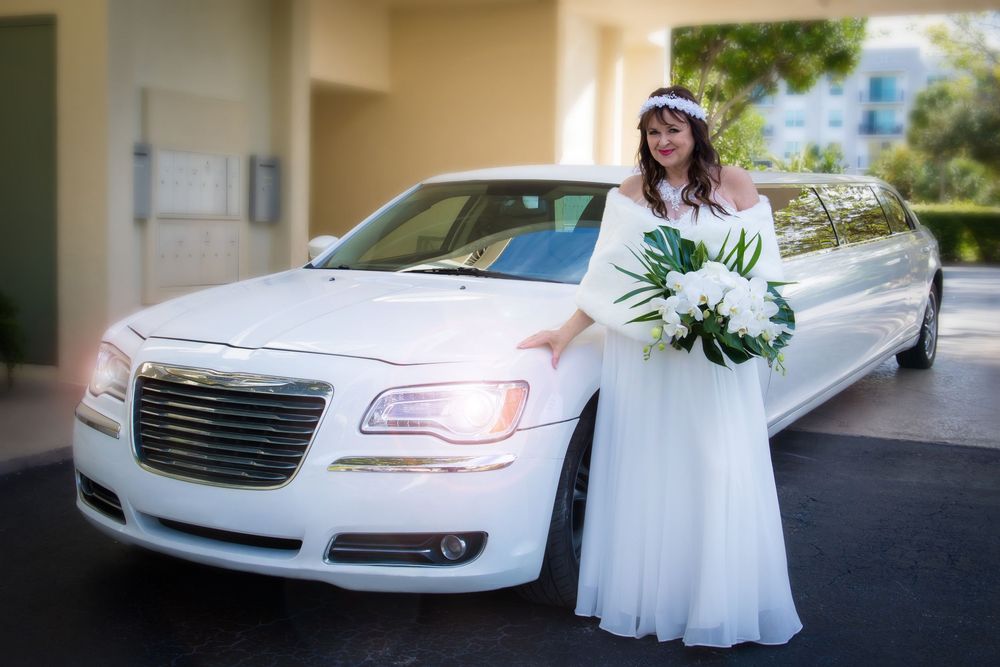 A white Chrysler limousine parked outside with someone in a flowing white wedding dress holding a floral bouquet.
